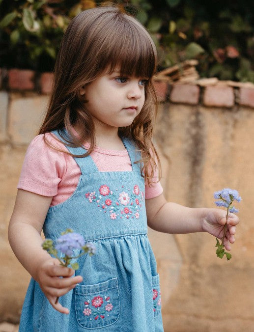 Young girl in a denim dress with floral embroidery holding flowers outdoors.