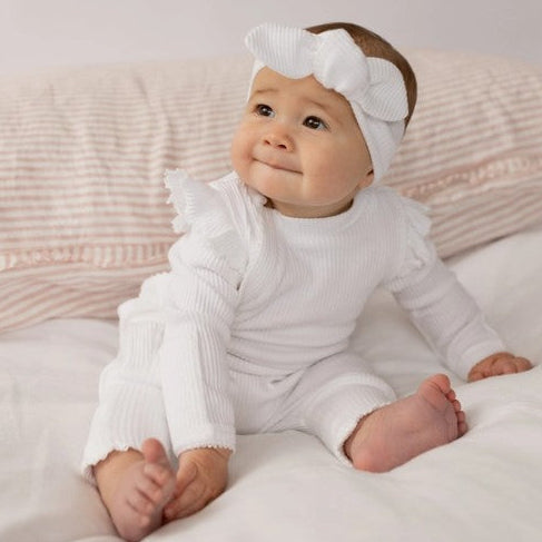 Baby in a white outfit with a headband sitting on a soft surface