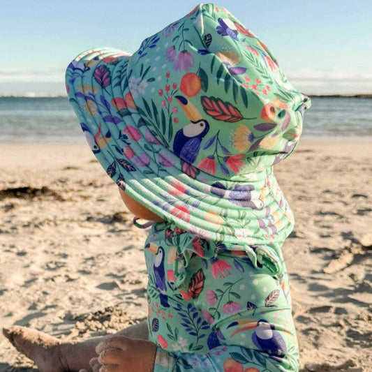 Child wearing a colorful floral and toucan patterned sun hat and outfit on a beach.